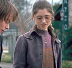 A young woman with light brown hair smiles while wearing the Stranger Things S01 Nancy Wheeler Purple Jacket by Zing Apparel, paired with a striped sweater and gold pendant necklace. Outdoors, a young man with brown hair is partly visible beside her.